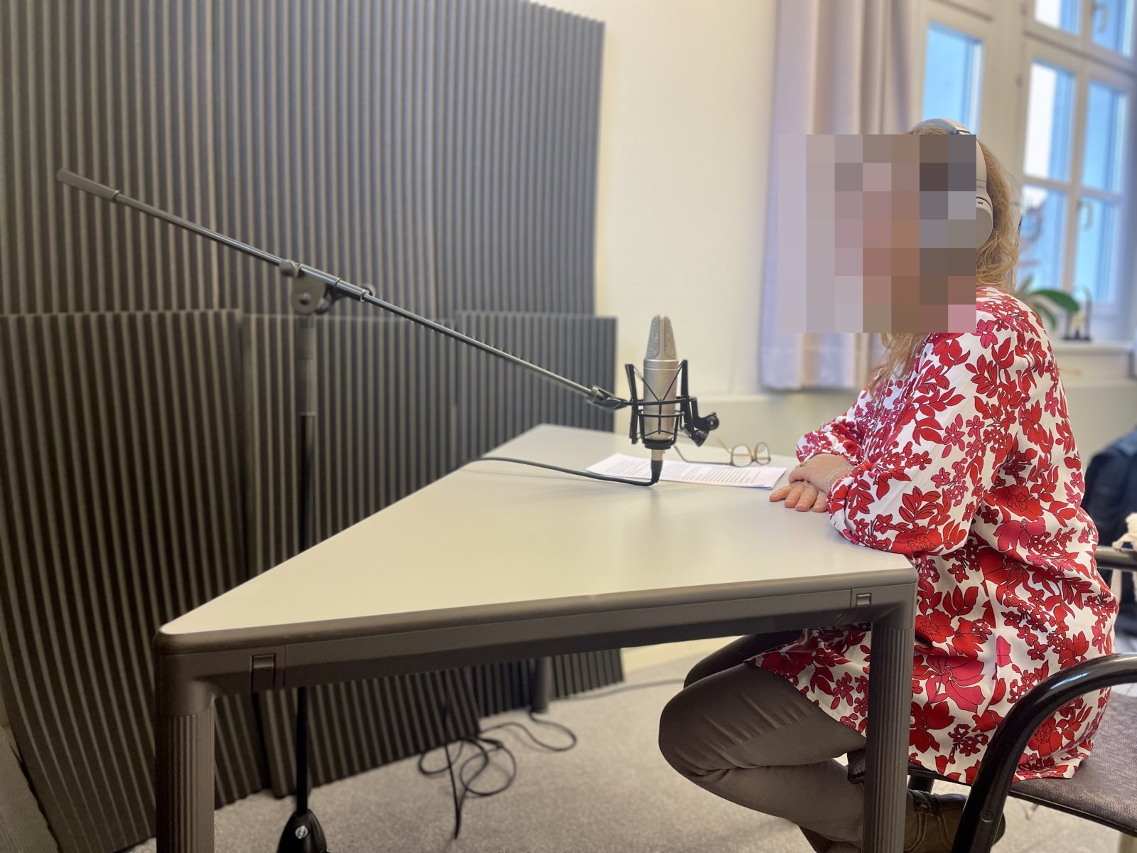 Eine Person sitzt am Mikrofon im Aufnahmeraum, vor sich Notizen; das Gesicht ist unkenntlich gemacht