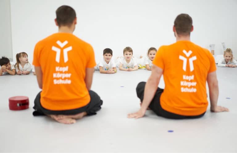Zwei Trainer in orangen Shirts sitzen vor einer Reihe Kinder, die Wortmarke gestapelt auf dem Rücken
